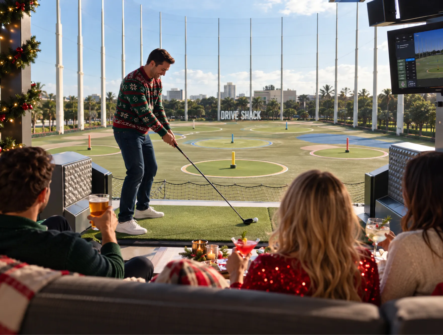 A golfer in a holiday sweater swings from a Drive Shack bay while coworkers cheer from behind the mat.